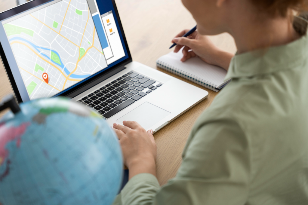 A woman is sitting at a table next to a globe, writing something in a notebook, and looking at a laptop screen with a map. 